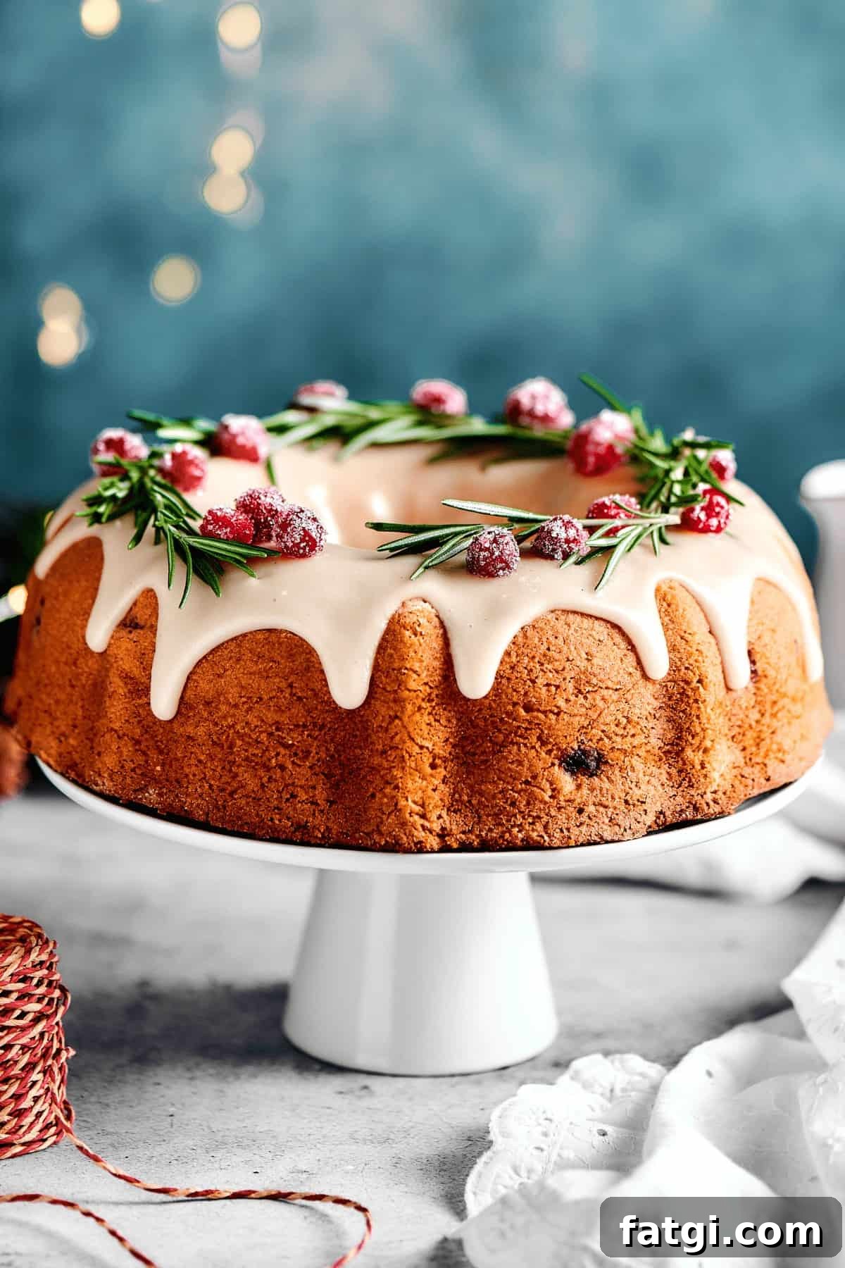 A bundt cake on a white cake stand decorated with white icing, sugared cranberries and rosemary sprigs.