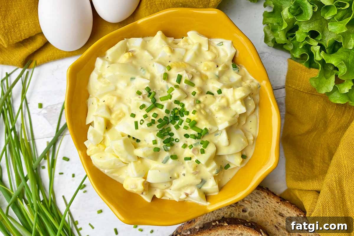 Horizontal top-down image of a yellow bowl filled with a creamy dish with chopped whites and yolks topped with chives next to bread slices and a yellow towel.