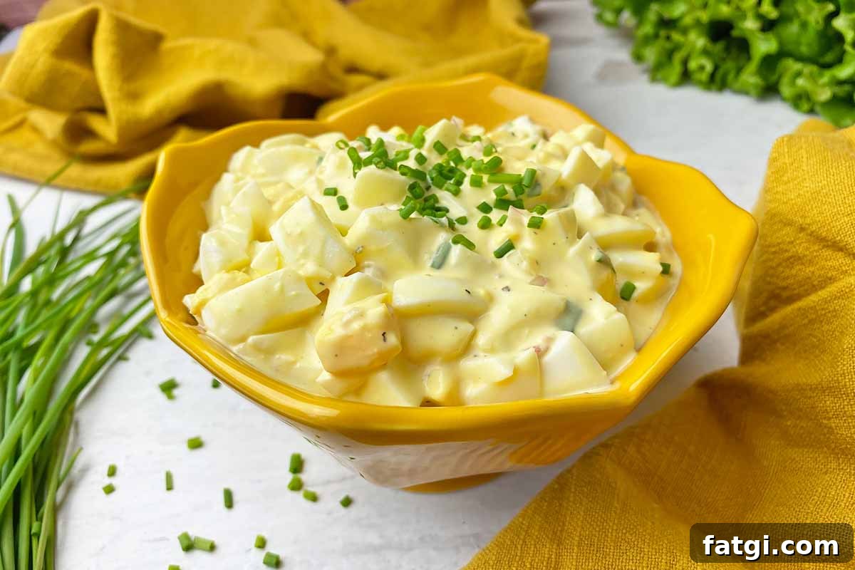 Horizontal image of a creamy, mayo-based mix of roughly chopped hard-boiled yolks and whites in a bright yellow bowl with a chive garnish.