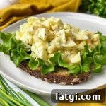 Horizontal image of an open-faced sandwich topped with lettuce and a creamy mix of hard-boiled yolks and whites on a white plate next to chives and a yellow towel.