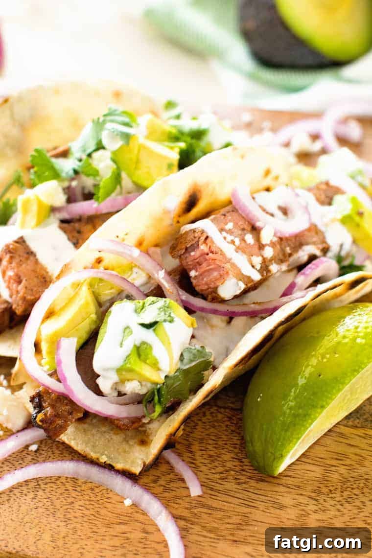 Flank steak tacos lined up on a wooden curling board topped with cilantro, red onion, avocado and a squeeze of lime.