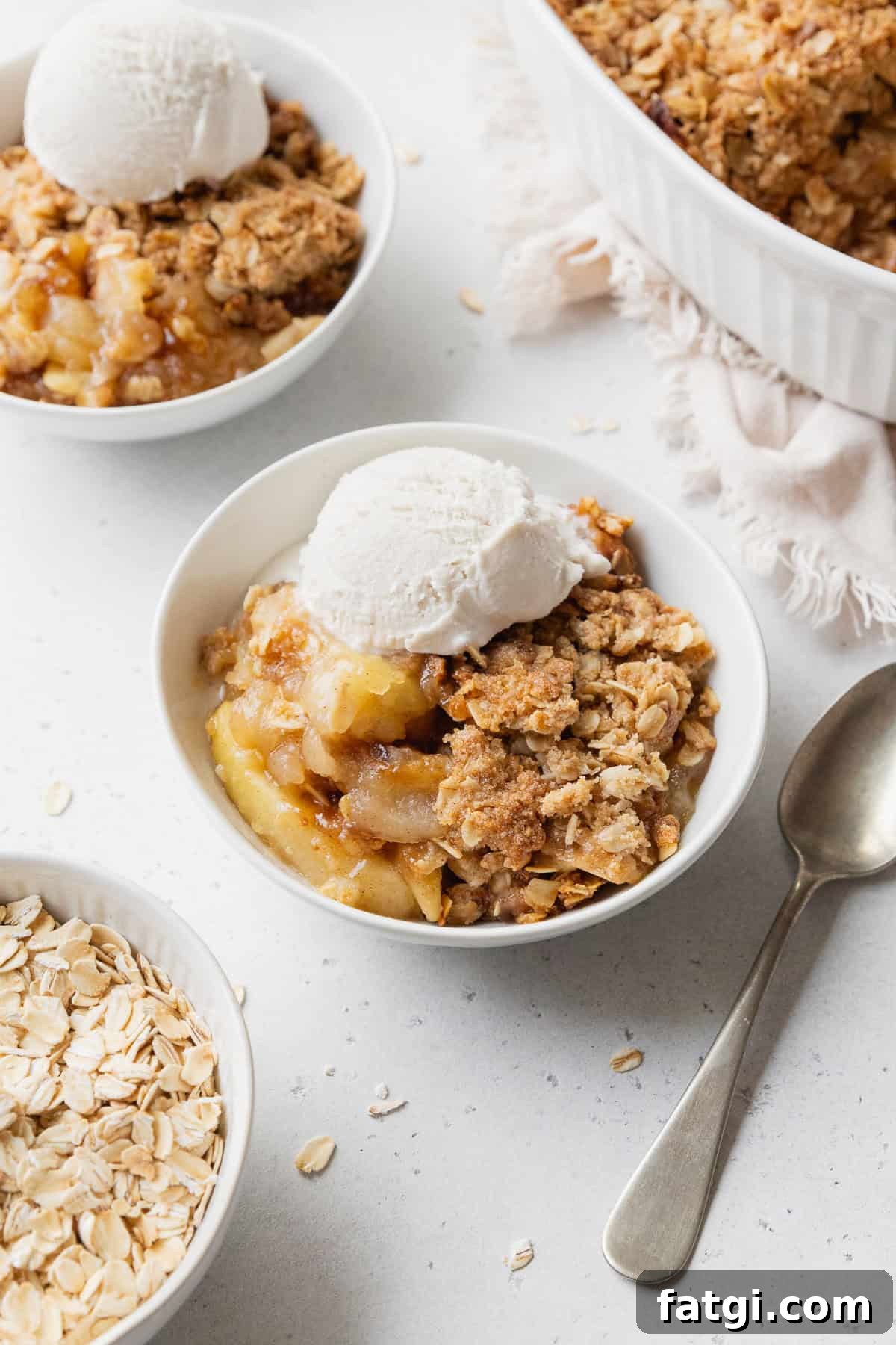 Homemade Gluten-Free Apple Crisp Recipe 2 Gluten-free apple crisp with vanilla ice cream in a small white bowl next to a spoon with more bowls placed nearby.