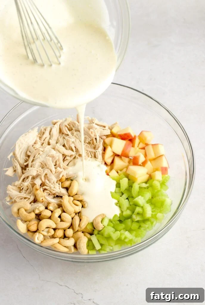 Dressing being poured over chicken, apples, cashews, and celery in a mixing bowl.