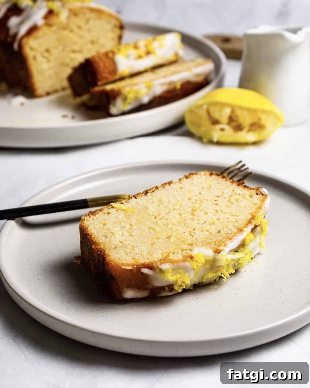 Gluten-Free Lemon Drizzle Loaf Cake Recipe 3 A slice of gluten-free lemon drizzle cake on a plate. There is a fork behind the cake. In the background is the rest of the loaf, sauce jug and half a squeezed lemon.
