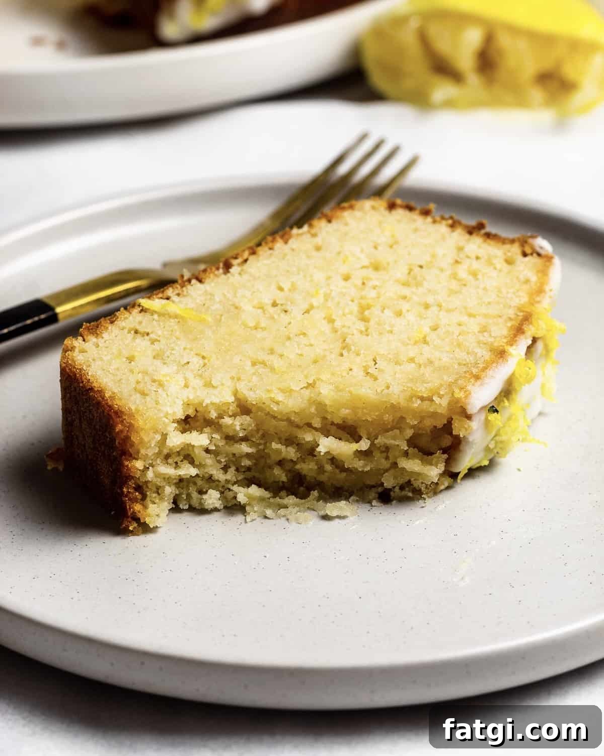 Gluten-Free Lemon Drizzle Loaf Cake Recipe 8 A half eaten slice of fluffy lemon drizzle cake on a plate, there is a fork and lemon in the background.
