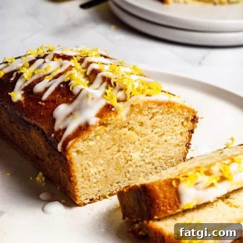 Gluten-Free Lemon Drizzle Loaf Cake Recipe 10 A gluten-free lemon loaf on a white plate, two slices have been cut and are lying off-centre. There are two plates stacked on top of each other in the background with a half eaten slice of cake.