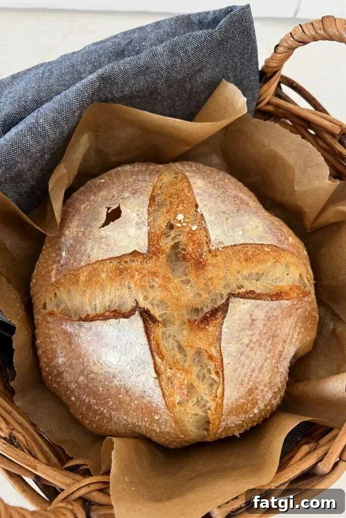 A sourdough bread bowl with a simple cross slashed into the top. It has been baked and is sitting inside a rattan basket lined with a denim blue dish towel.