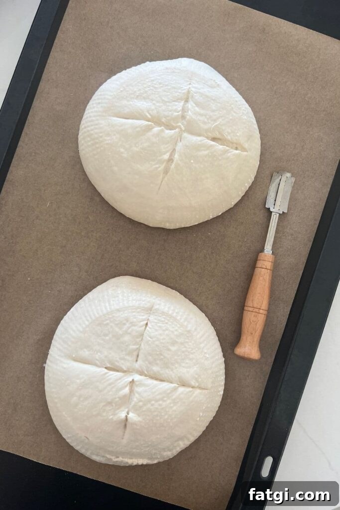 Two loaves of sourdough bread sitting on a piece of parchment paper. They have been scored with a cross on the top. There is a wooden handled lame sitting to the right of the bread bowls.