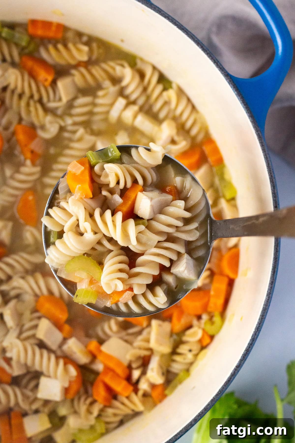 A ladle filled with rotini pasta, diced carrots, celery, and pieces of turkey is held above a pot containing more of the same soup. The pot has a blue handle, and fresh celery is visible on the side.