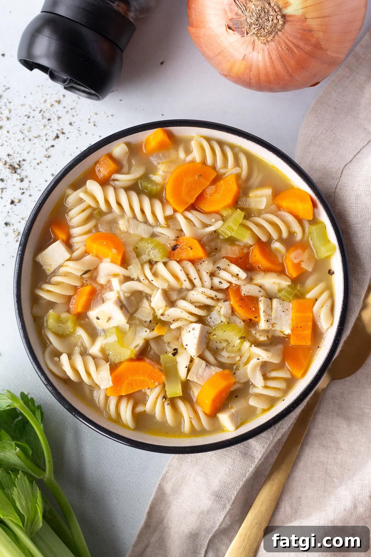 A bowl of pasta soup with fusilli, diced carrots, celery, and turkey pieces, garnished with black pepper. An onion and a celery stalk are nearby, along with a black pepper grinder. A spoon is placed on a beige napkin beside the bowl.