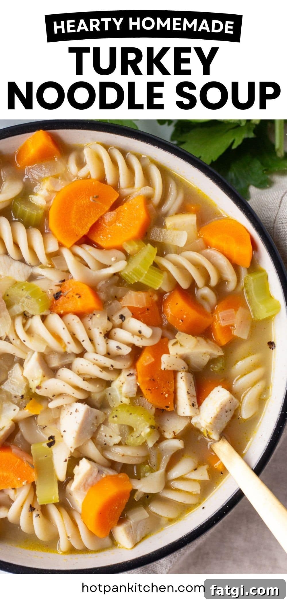 A bowl of turkey noodle soup filled with spiral pasta, chunks of turkey, sliced carrots, and celery. The soup is topped with black pepper. A wooden spoon rests in the bowl. Text on top reads "Hearty Homemade Turkey Noodle Soup.