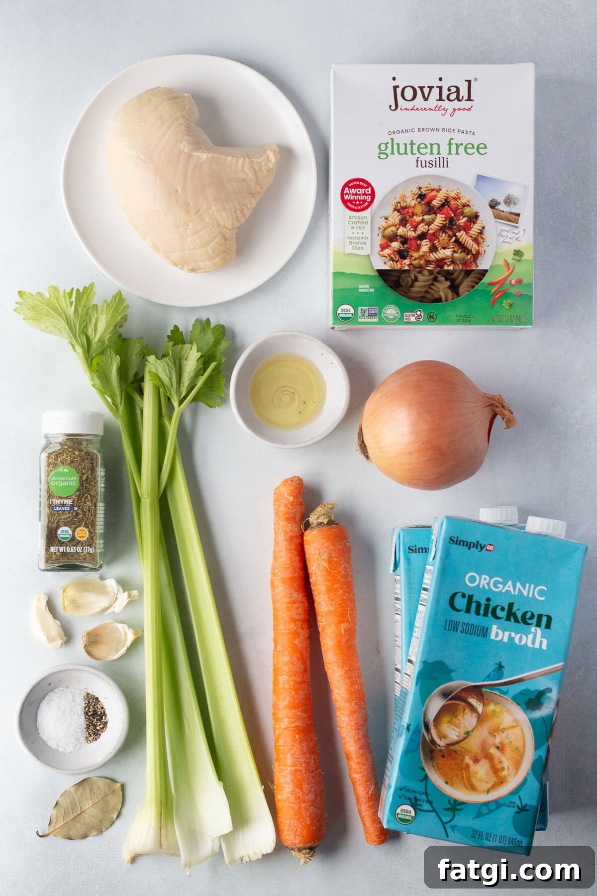 A collection of ingredients for a recipe: cooked turkey breast, gluten-free fusilli, celery stalks, an onion, carrots, chicken broth, dried thyme, oil, garlic cloves, salt, and pepper on a light surface.