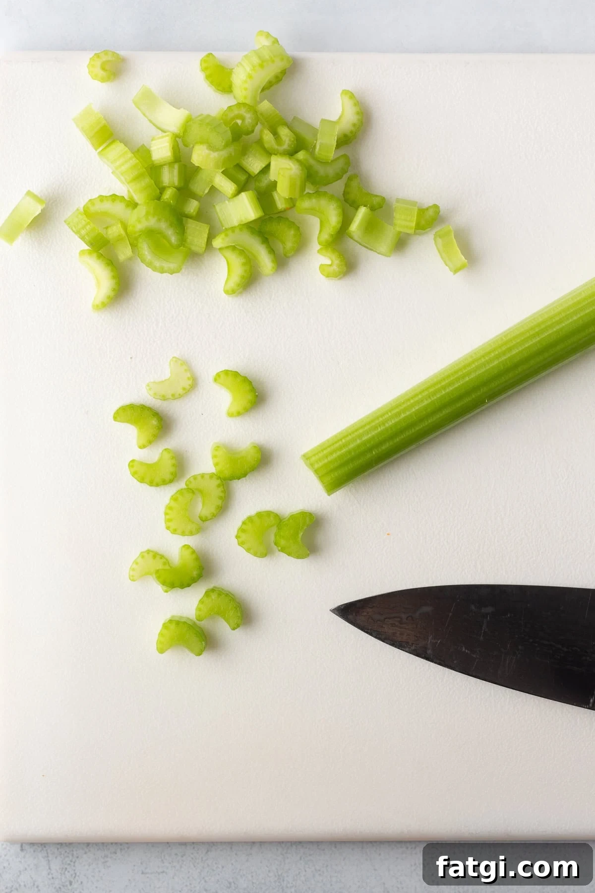 Chopped celery pieces and a whole celery stalk on a white cutting board with a black knife.