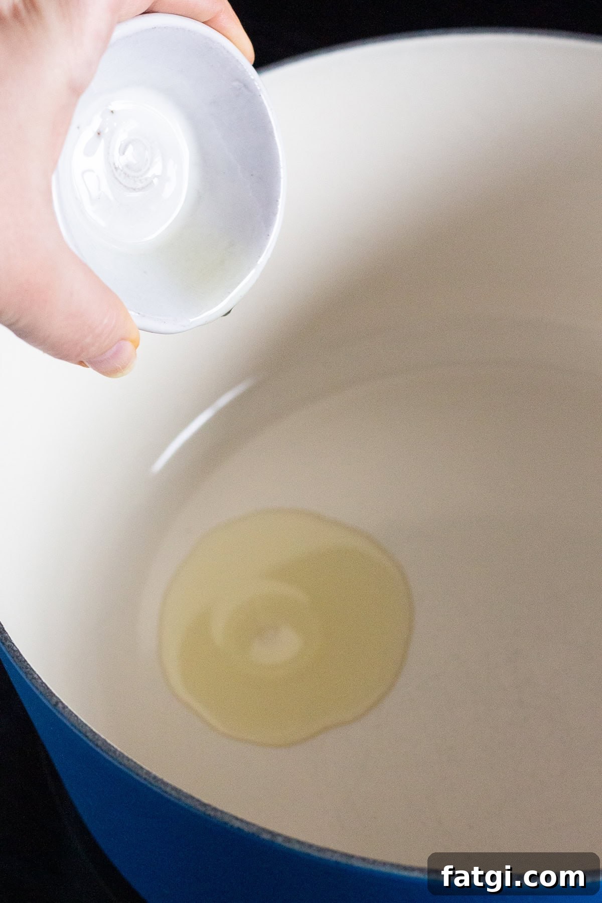 A person pours a small amount of oil from a white container into a large blue pot with a white interior. The pot sits on a stovetop.