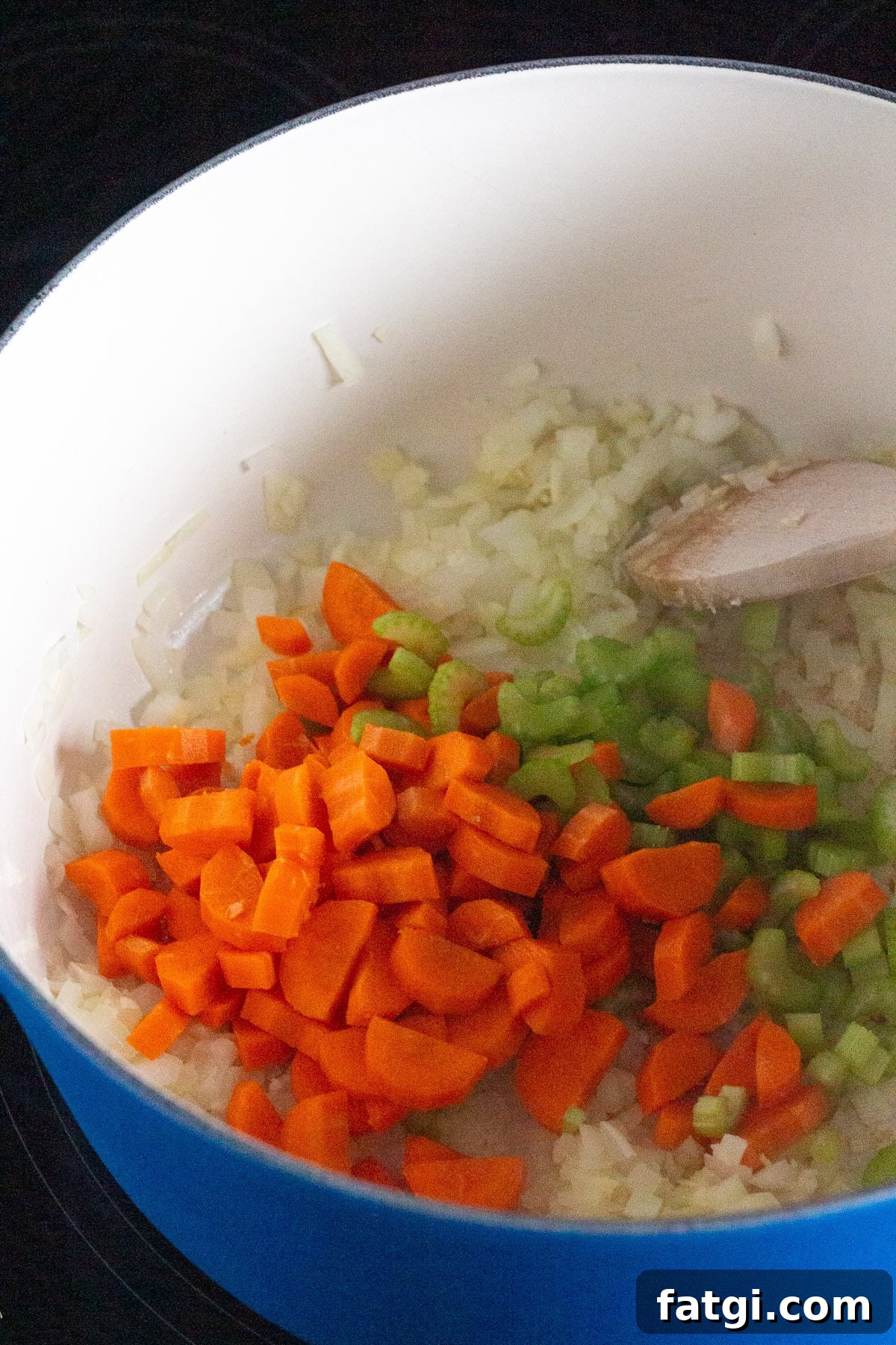 A blue pot on a stove contains chopped onions, carrots, and celery being sautéed with a wooden spoon. The vegetables are in the early stages of cooking.