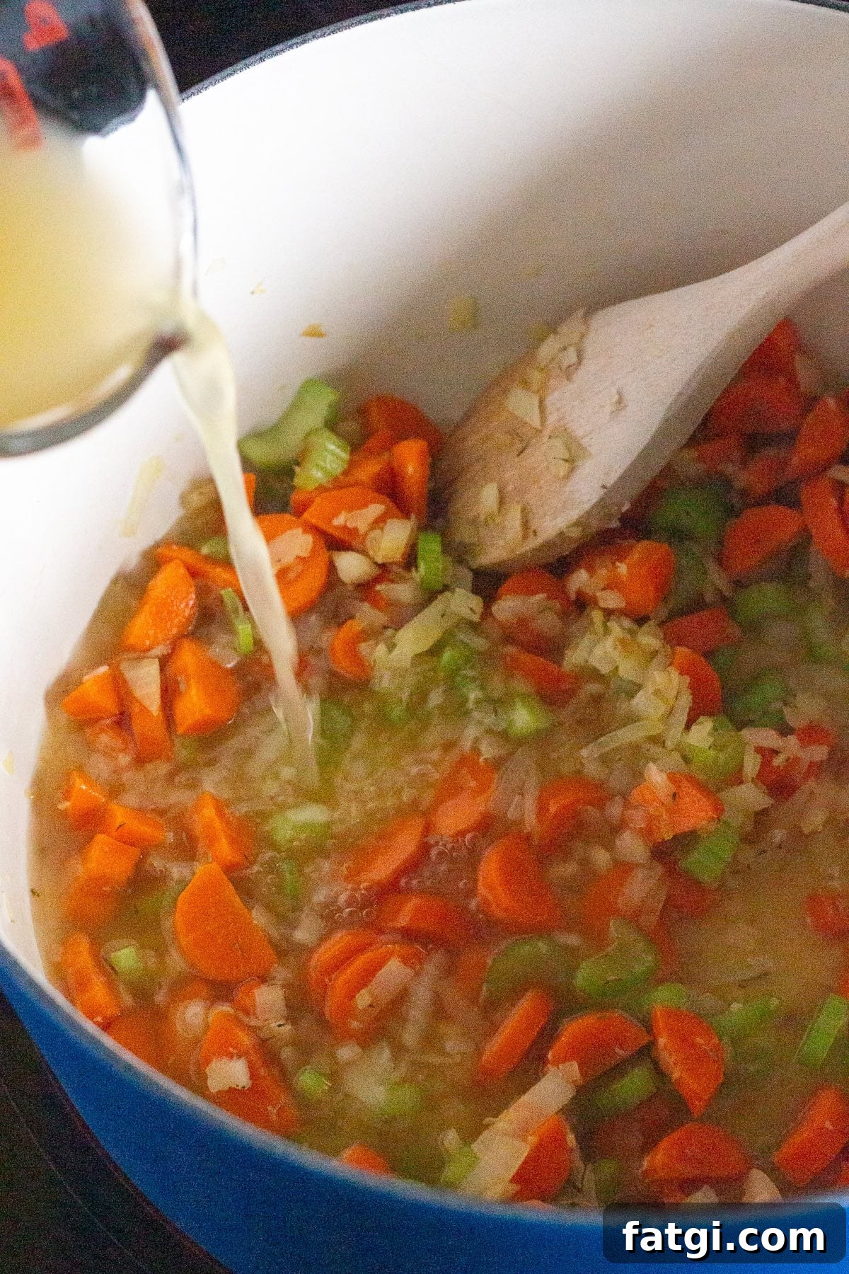 Chopped carrots, celery, and onions are being cooked in a pot. A wooden spoon is stirring the mixture, and broth is being poured from a measuring cup into the pot.