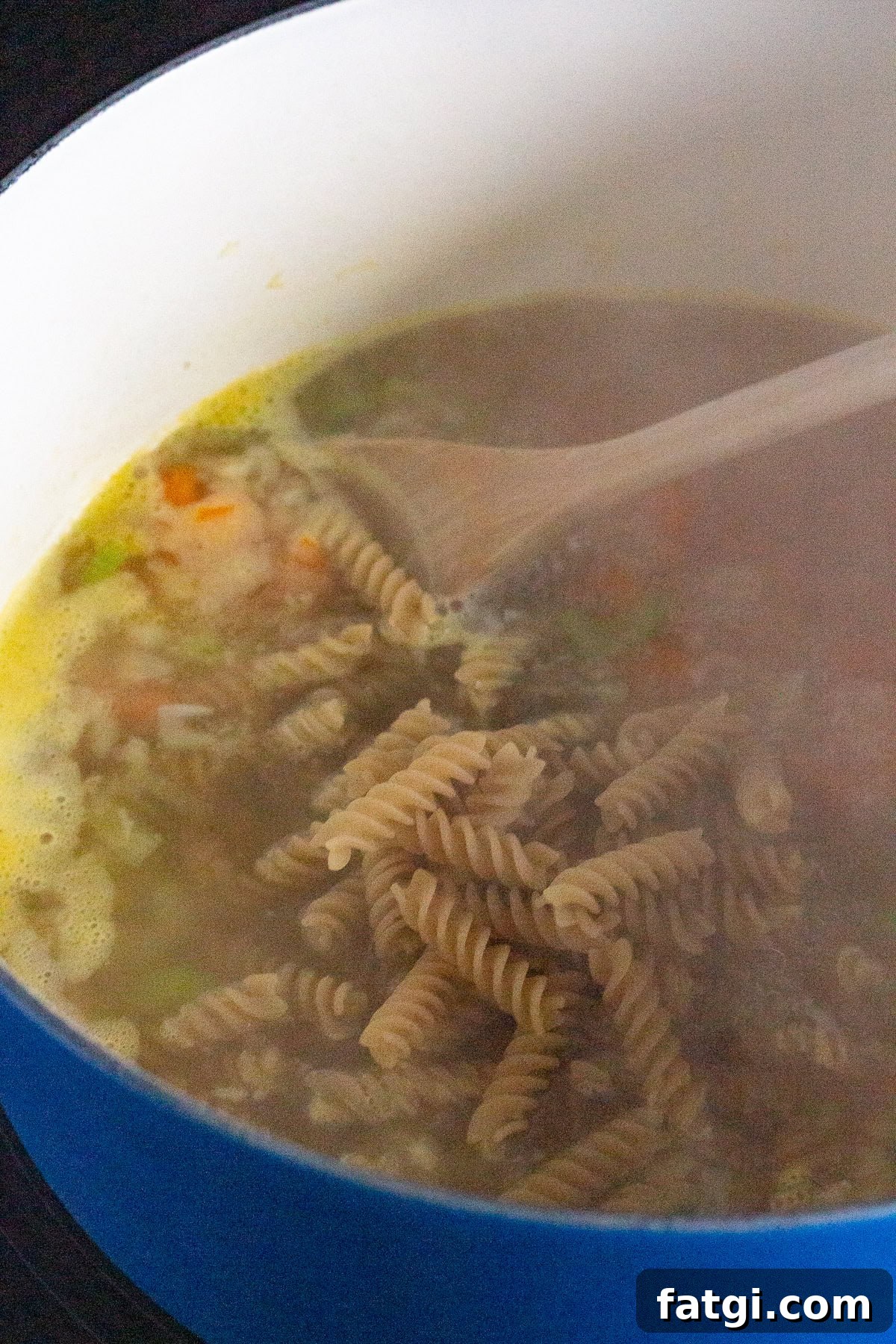 A blue pot on a stove contains a simmering soup with rotini pasta, diced vegetables, and broth. A wooden spoon is visible in the mixture. Steam rises from the pot, indicating heat.