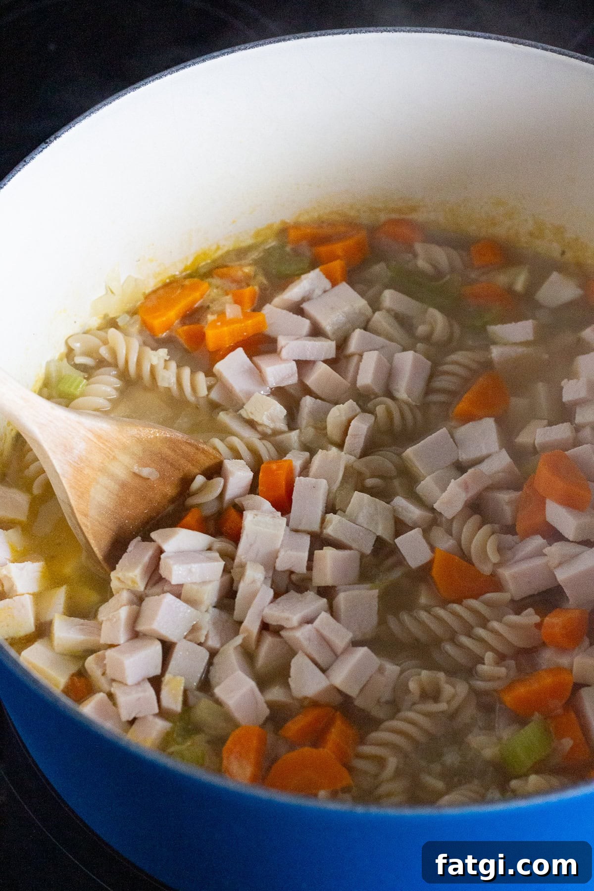 A blue pot containing a soup mixture with spiral pasta, chopped carrots, celery, and cubes of turkey is being stirred with a wooden spoon. Steam rises from the pot, indicating the soup is hot.