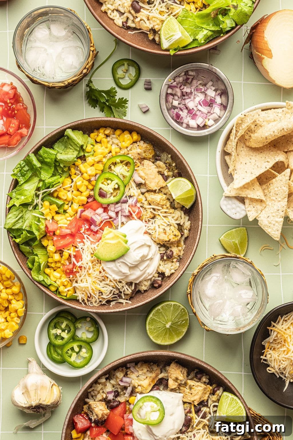 A bowl with white rice, chicken, black beans, cheese, corn, lettuce, tomatoes, diced onions, sour cream, lime wedges, and sliced jalapeno. Surrounded by tortilla chips, bowl of diced red onion, bowl with sliced jalapeno, lemon wedges, and a glass of ice water.