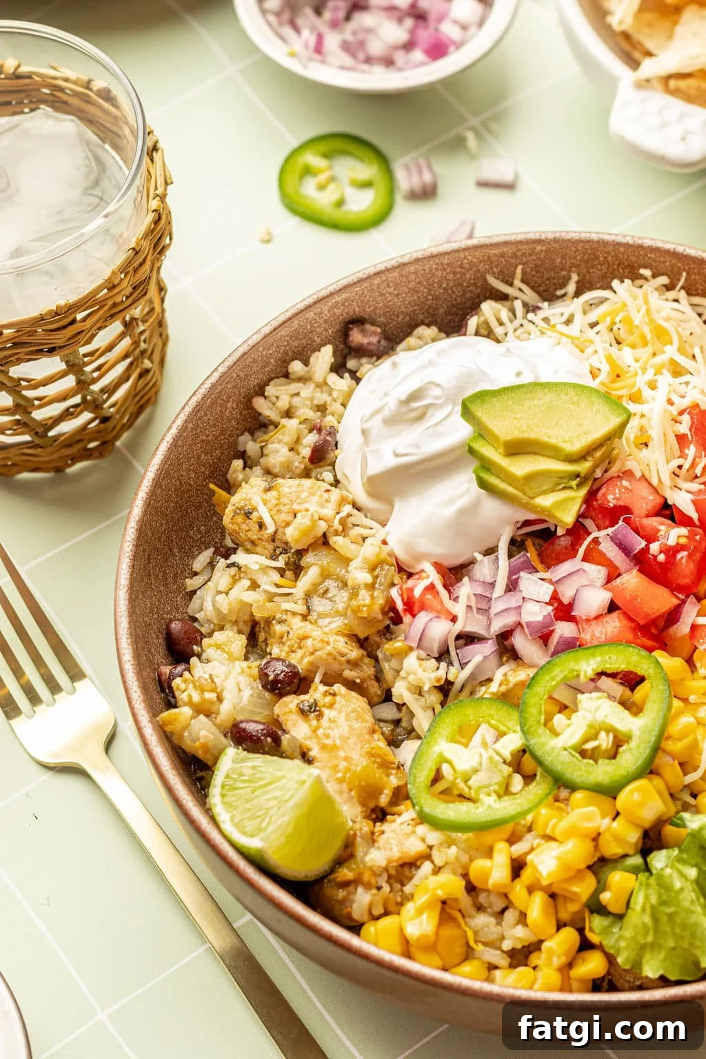 Close up image of a bowl with chicken, rice, sour cream, shredded cheese, lettuce, tomatoes, avocado, sliced jalapeno, and lime wedges.