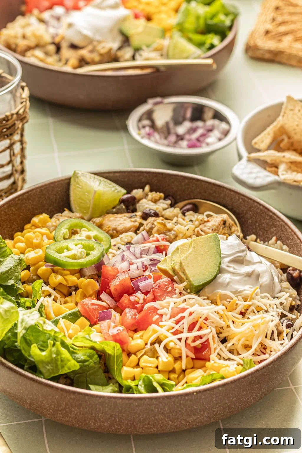 Close up image of a bowl with chicken, rice, sour cream, shredded cheese, lettuce, tomatoes, avocado, sliced jalapeno, and lime wedges.