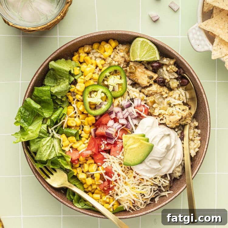 A bowl with a gold fork and spoon and white rice, chicken, black beans, cheese, corn, lettuce, tomatoes, diced onions, sour cream, lime wedges, and sliced jalapeno.