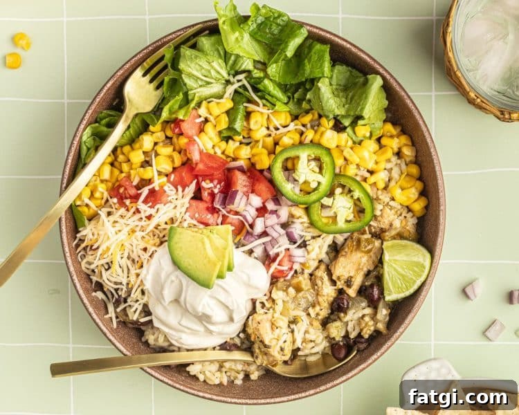 A bowl with a gold fork and spoon and white rice, chicken, black beans, cheese, corn, lettuce, tomatoes, diced onions, sour cream, lime wedges, and sliced jalapeno.