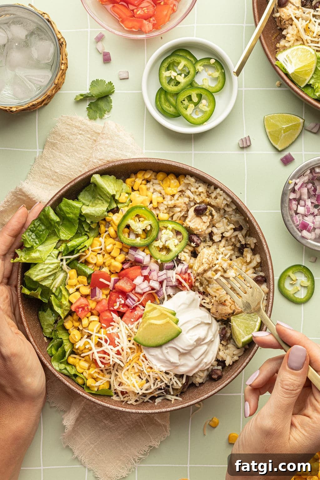 A bowl with a gold fork and spoon and white rice, chicken, black beans, cheese, corn, lettuce, tomatoes, diced onions, sour cream, lime wedges, and sliced jalapeno.