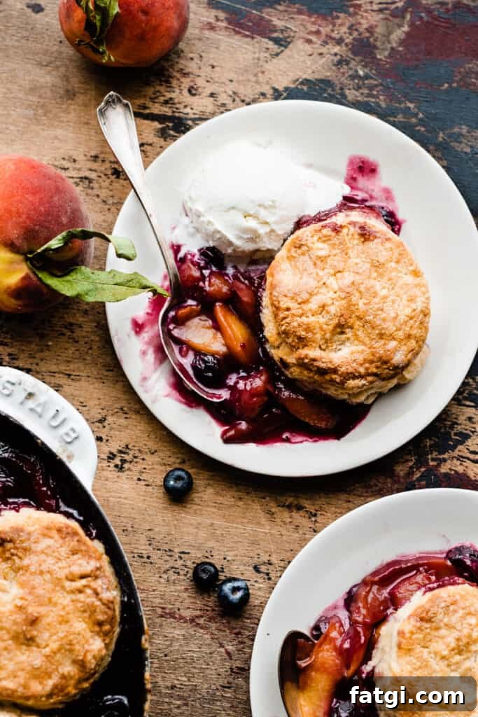 Two plates of the vibrant blueberry peach cobbler, with fresh blueberries and peaches scattered around, on a wooden surface.