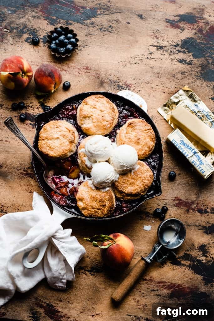 Blueberry Peach Cobbler in a cast iron skillet, topped with biscuits and vanilla ice cream, on a wooden surface.