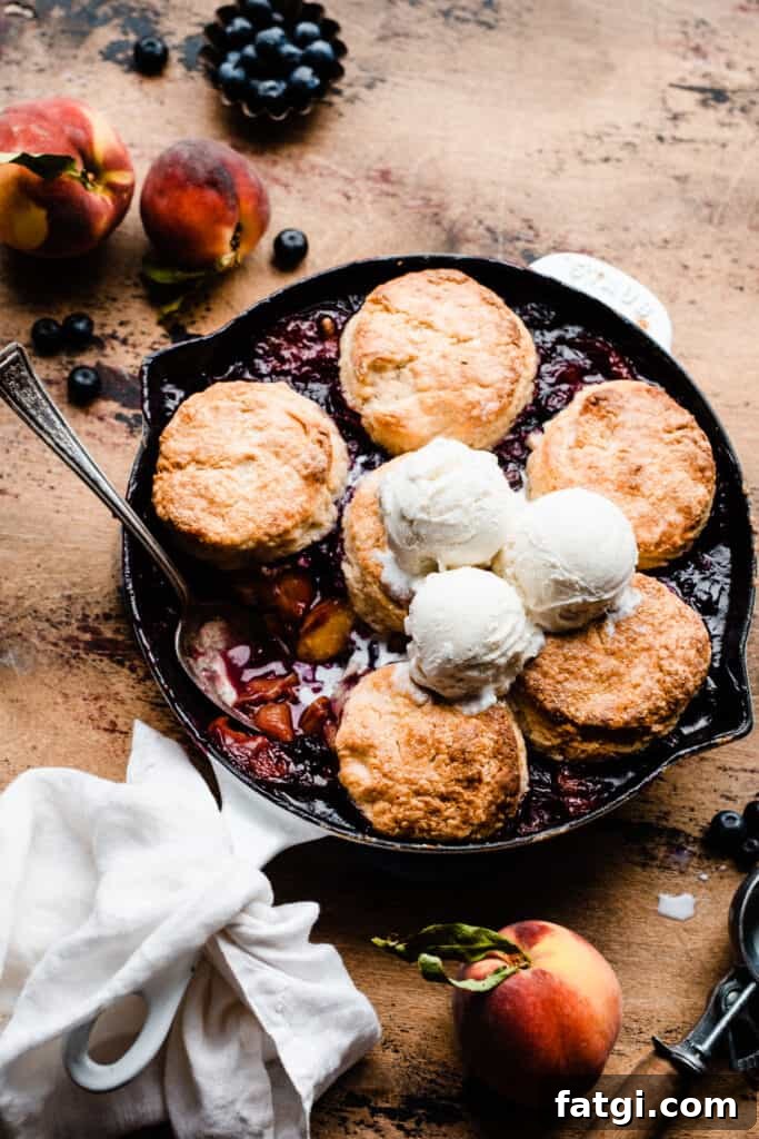 A cast iron skillet of blueberry peach cobbler, topped with golden brown biscuits and scoops of vanilla ice cream.