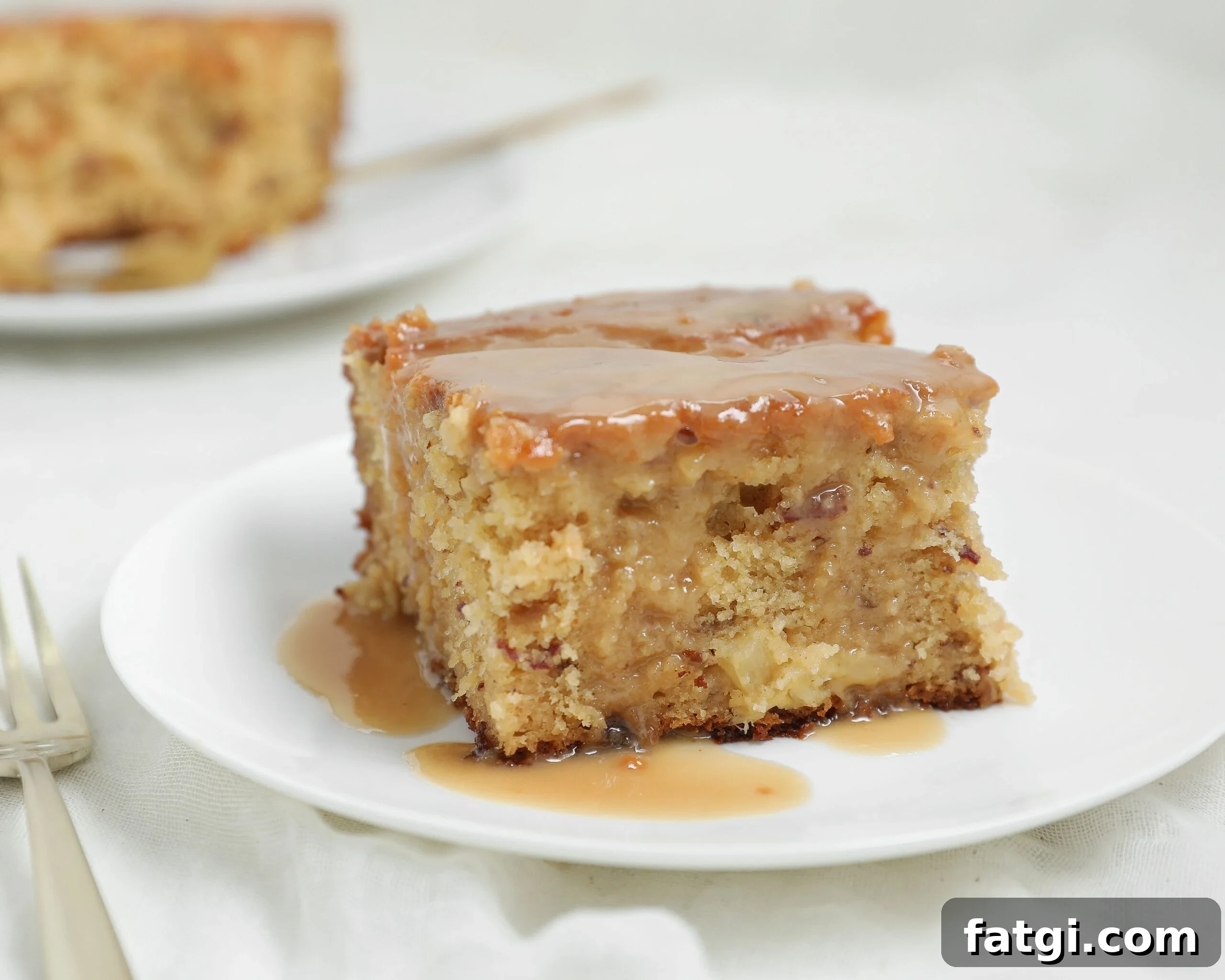 A slice of moist Pineapple Sticky Toffee Pudding Cake with luscious pineapple toffee sauce on top sits on a white plate, with a fork nearby. Another slice of cake is visible in the blurred background.