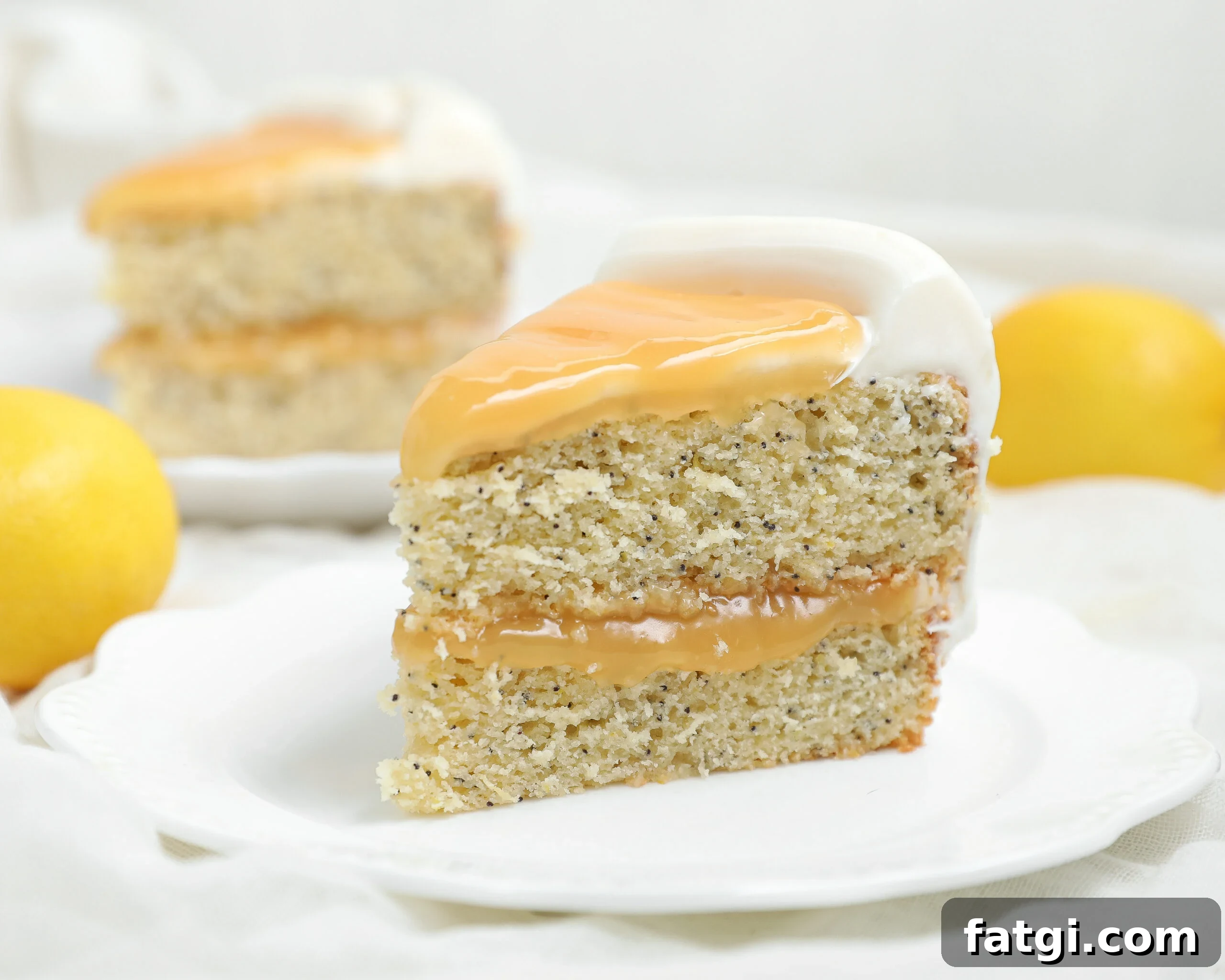A slice of Lemon Poppy Seed Cake with creamy white frosting and yellow glaze sits on a white plate, surrounded by whole lemons and another tempting slice in the background.