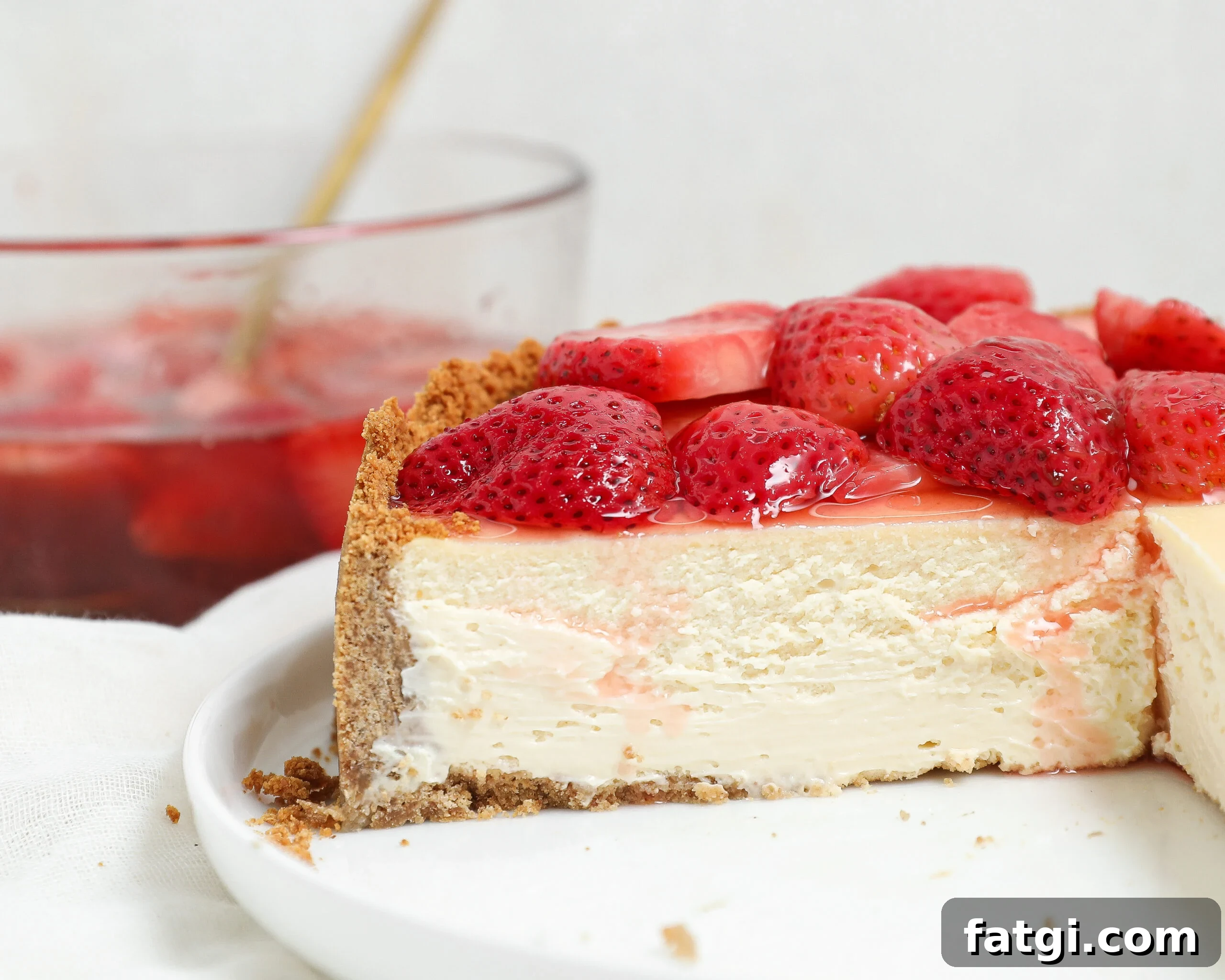 A close-up of a cheesecake with a graham cracker crust, topped with fresh strawberries. A slice is missing, showing the creamy texture inside. In the background, there is a glass bowl with more strawberries.