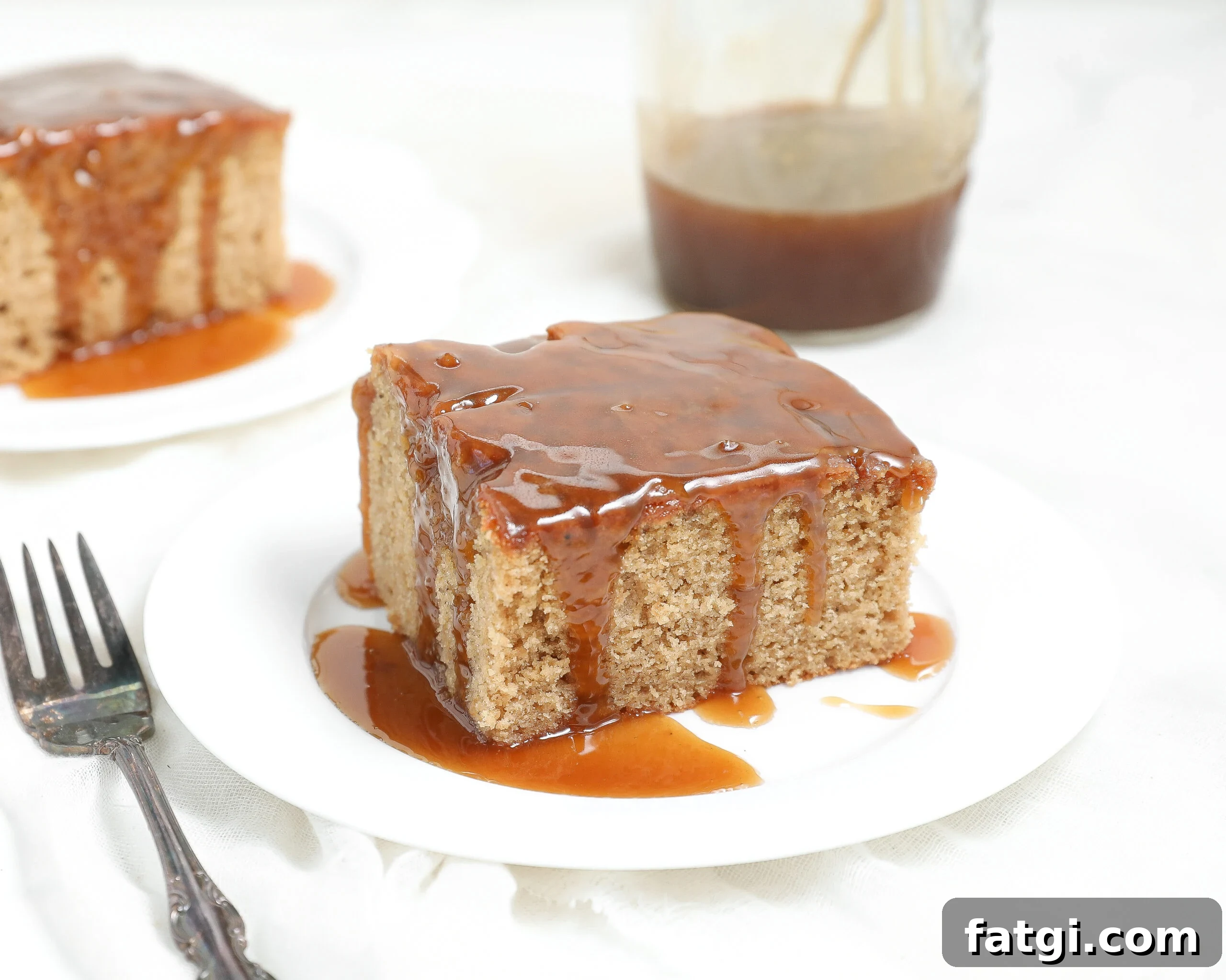 A square slice of moist biscoff sticky pudding cake covered in caramel sauce sits on a white plate, with a fork beside it and a jar of caramel sauce in the background. Another slice of cake is visible in the background.