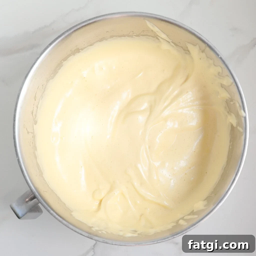 A metal mixing bowl filled with thick, pale yellow sponge cake batter or whipped mixture sits on a white marble surface.