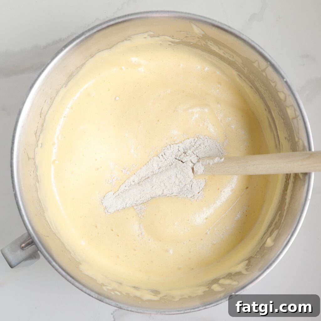 A metal mixing bowl filled with pale yellow sponge cake batter, with a heap of flour sitting on top. A wooden spoon is partially submerged in the mixture, resting on the side of the bowl.
