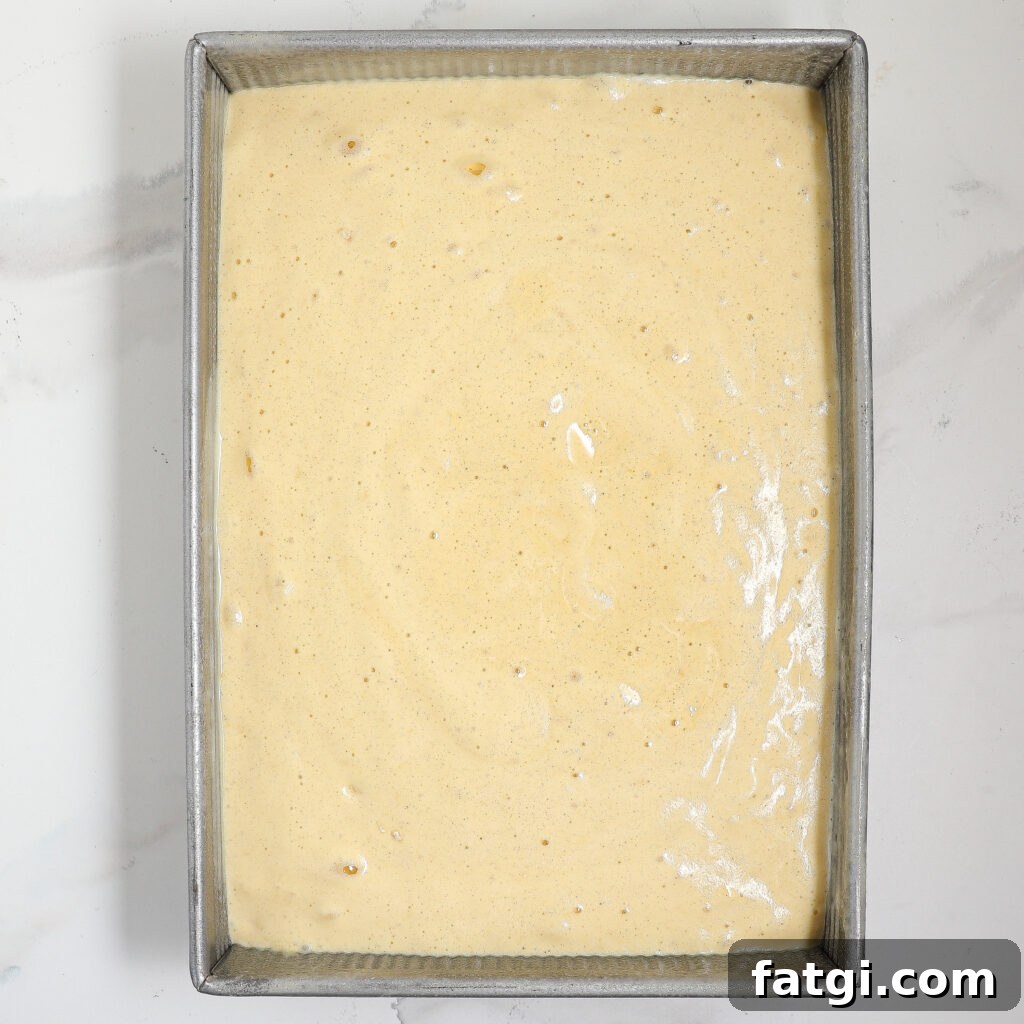A rectangular metal baking pan filled with an even layer of pale yellow sponge cake batter, set on a white marble surface.