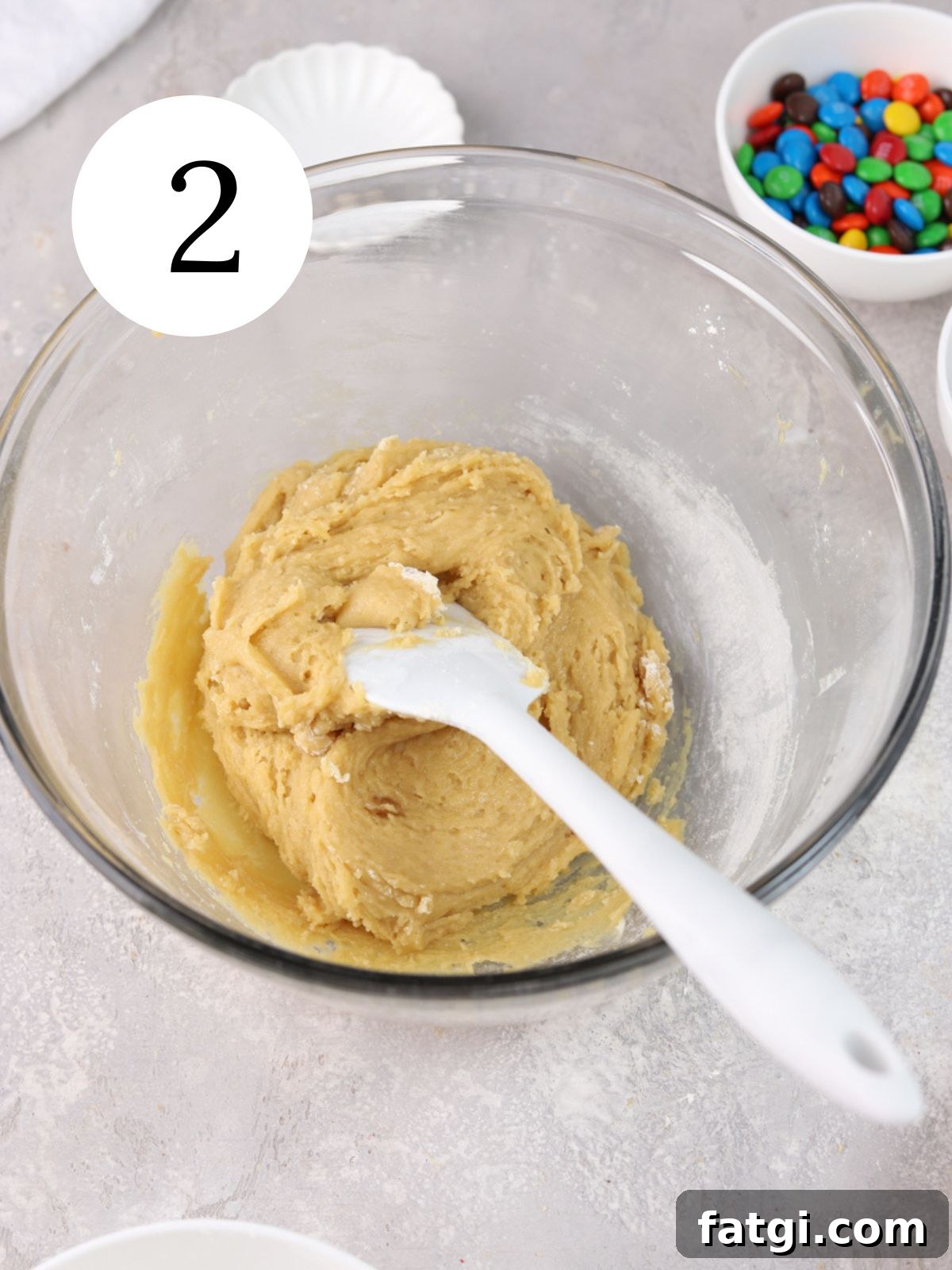 A mixing bowl with a spatula and cookie dough and a white bowl with M&M candy.