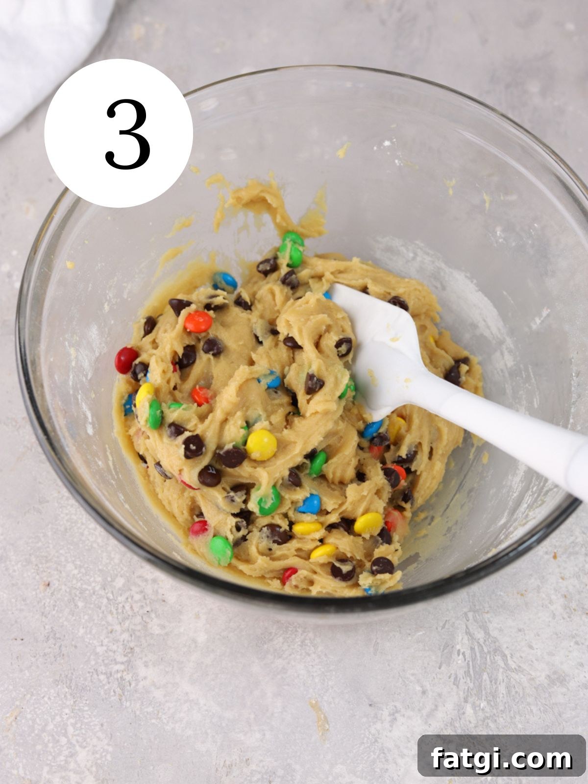 A glass mixing bowl with chocolate chip M&M cookie dough and a spatula.