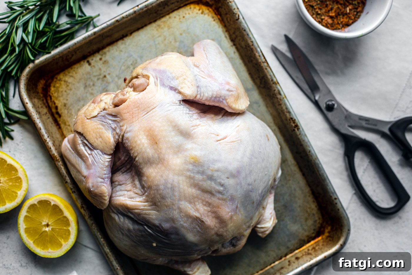 How to Spatchcock a Chicken: Step-by-Step Video 6 Baking sheet with raw whole chicken next to kitchen shears, seasoning, lemon halves, and fresh herbs.
