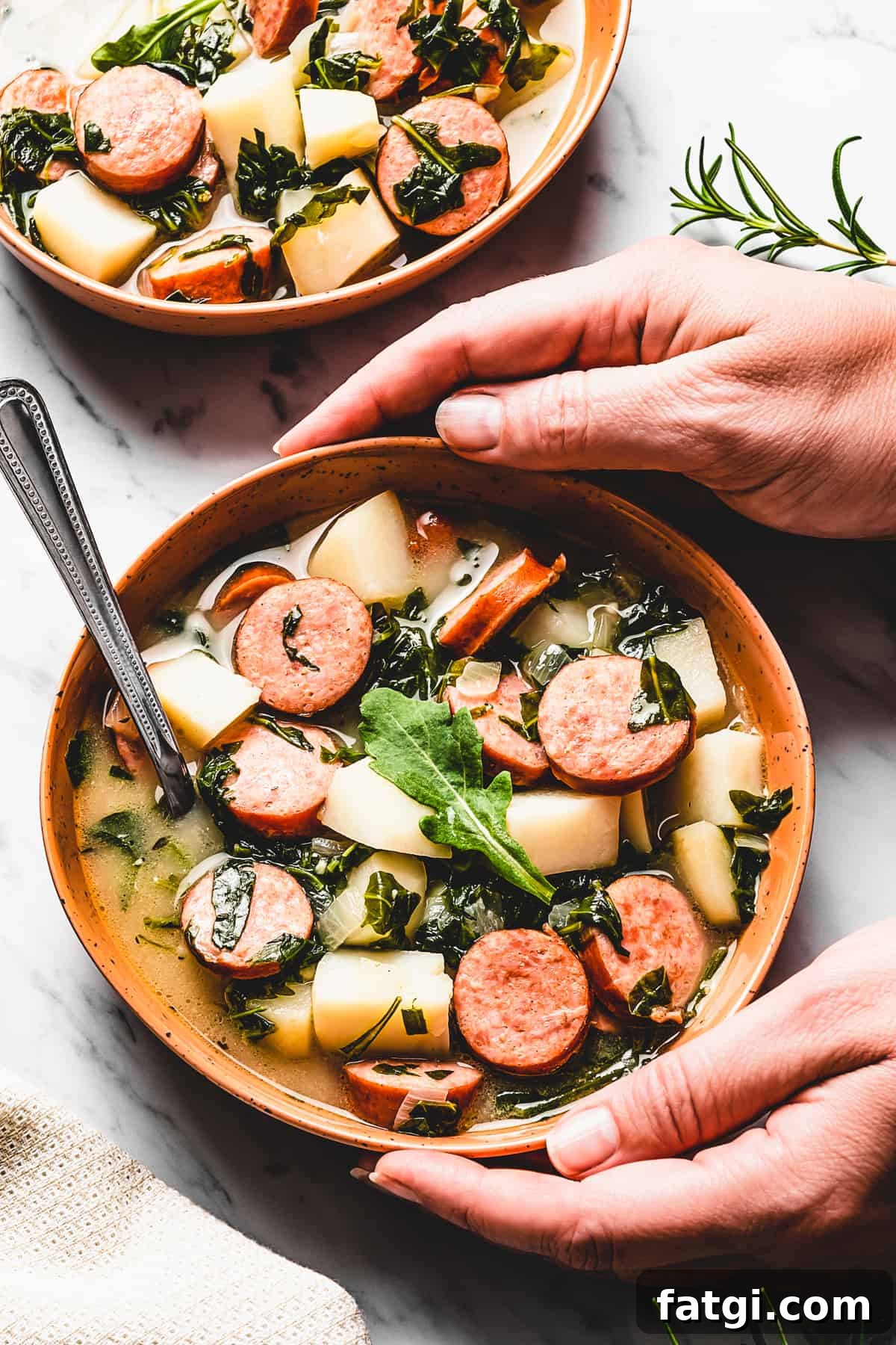 Hands picking up a bowl of potato soup with kale and sausages.