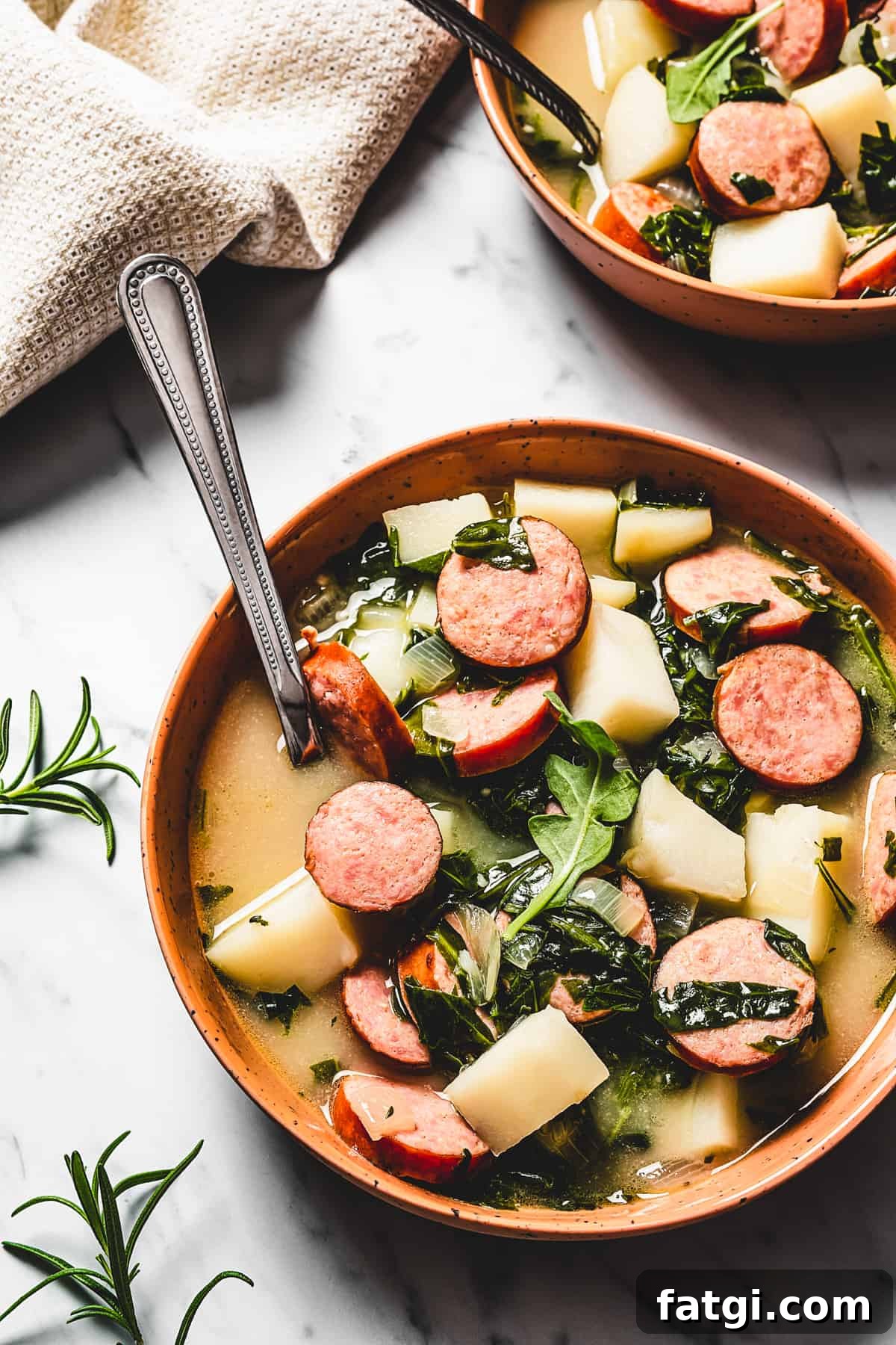 Bowls of easy potato and sausage soup on a table with herbs, salt, pepper, spoons, and a cloth napkin.