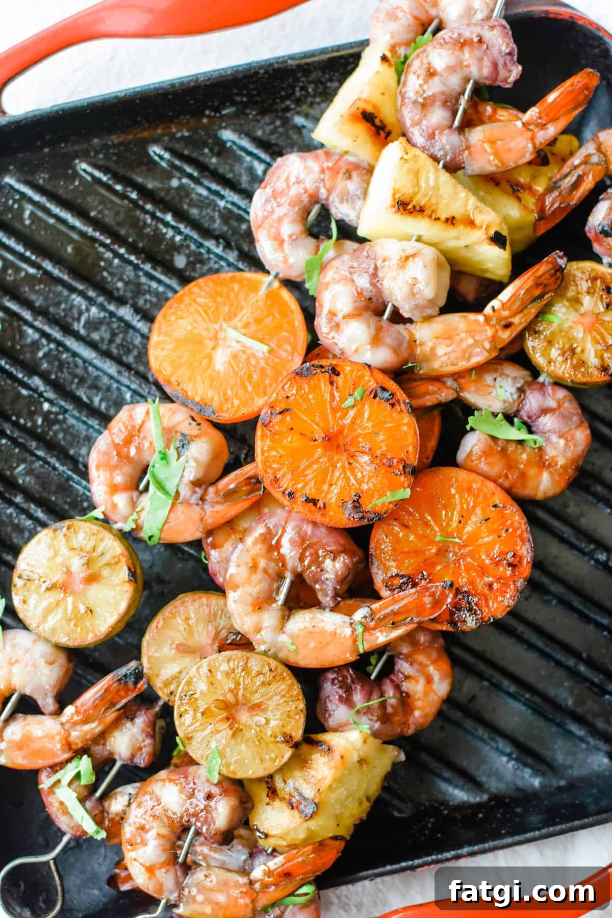 Grilled Shrimp Skewers with Sangria Marinade 5 Overhead image of grilled shrimp, tangerine halves, key limes, and pineapple on metal skewers sitting on a grill pan with red edges.