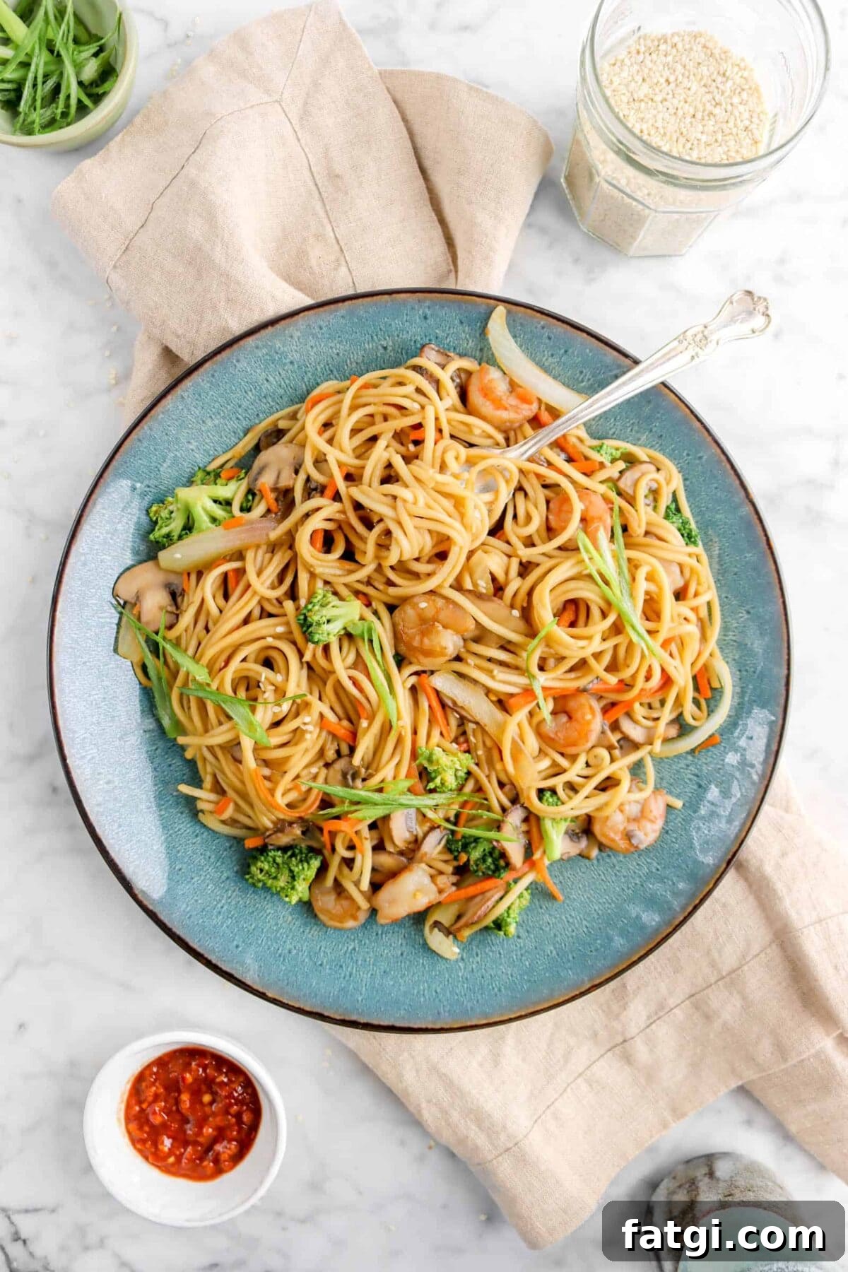 lo mein in blue plate on tan napkin with jar of sesame seeds, chili paste, and green onions