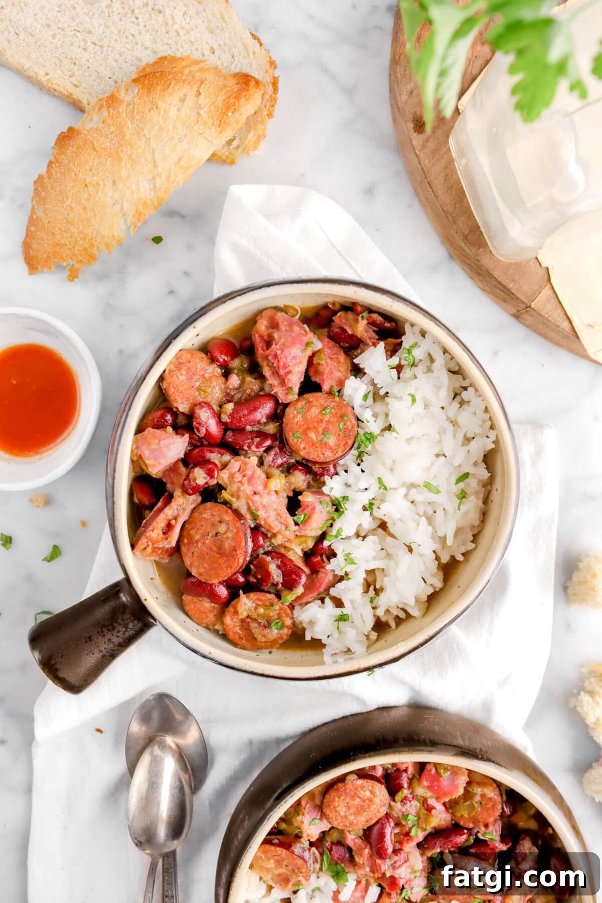 close up overhead shot of red beans and rice with bowl of hot sauce, bread chunk and two spoons with a wood serving board beside