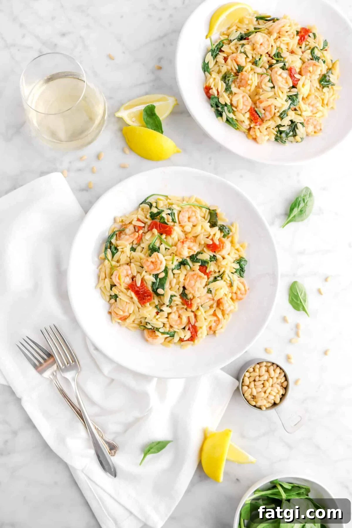 two bowls of shrimp orzo on marble counter with white napkin, two forks, spinach leaves, pine nuts, lemon wedges, and glass of wine