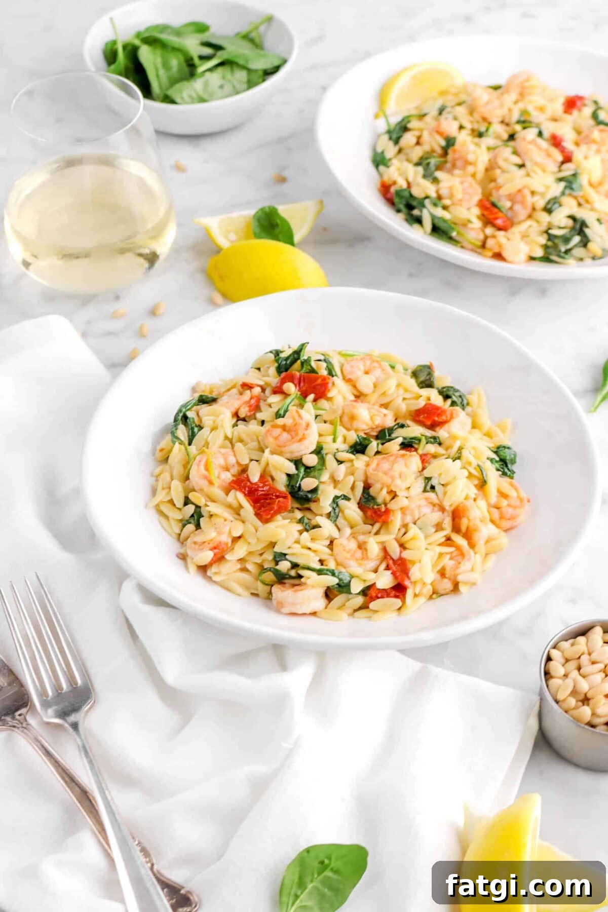 side shot of shrimp orzo in two bowls with glass of wine, lemon wedges, and bowl of spinach behind