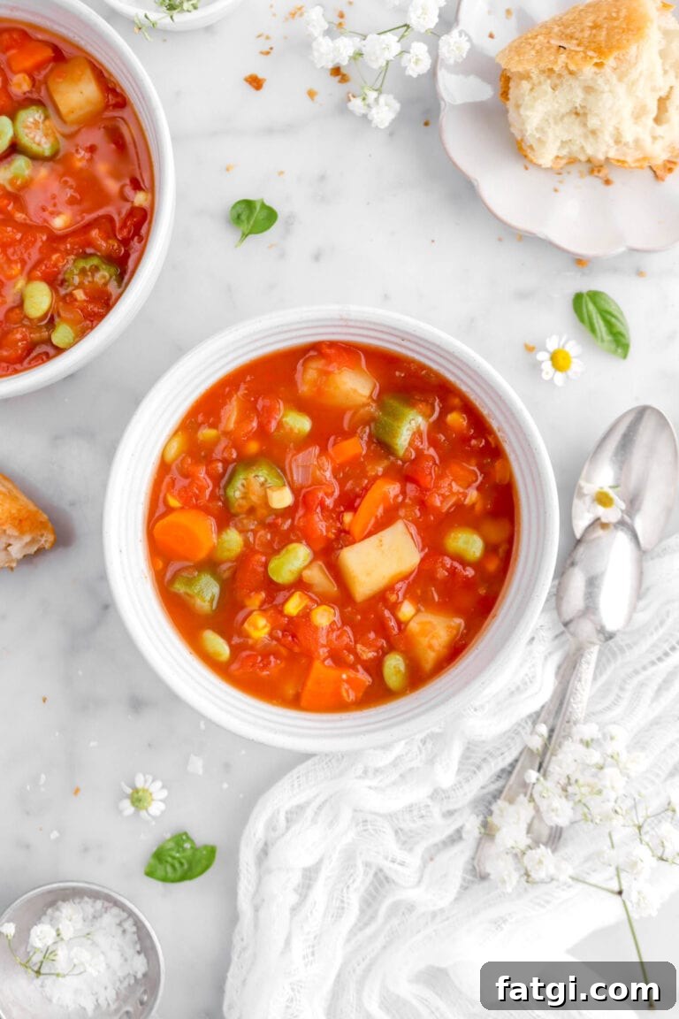 close up of bowl of vegetable soup on marble surface with another bowl of soup beside, chunk of bread, spoons, and flowers around.
