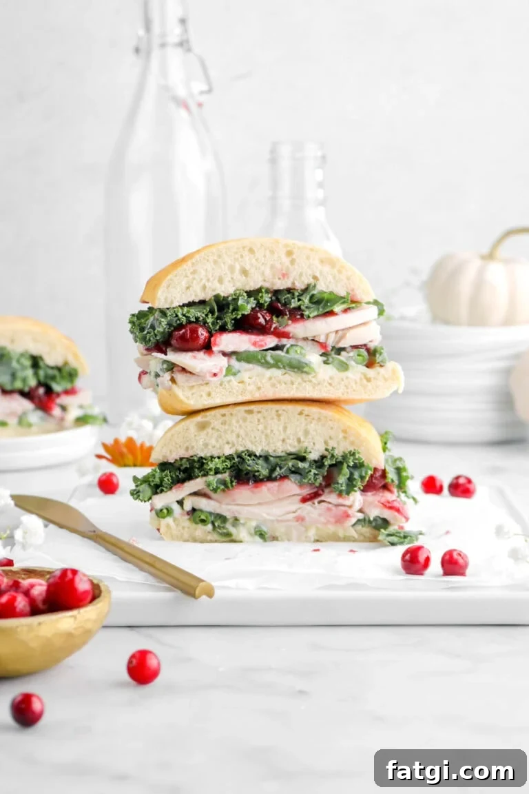 front shot of two halves of sandwich stacked on white tray with gold bowl of cranberries in front, stack of white plates behind with small white pumpkin on top.